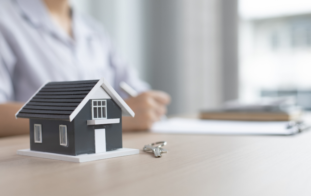 Miniature house model on a desk with a set of keys beside it, as a person writes in the background—evoking real estate or home buying.