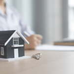 Miniature house model on a desk with a set of keys beside it, as a person writes in the background—evoking real estate or home buying.