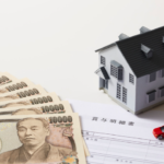 Fanned Japanese yen banknotes with a model house, a small red car, and a contract on a table—illustrating a loan or financing concept.