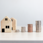 Two wooden house models sit beside progressively taller stacks of coins, illustrating home savings or real estate investment growth.