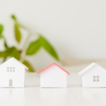 Three small white paper houses on a white table, with a blurred green plant in the background (middle roof is red).