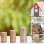 Coin stacks increasing in height from left to right beside a clear glass jar filled with coins, topped with a tiny toy house.