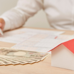 Person writing on documents at a desk, with a small paper house model and a fan of cash nearby, suggesting mortgage planning.