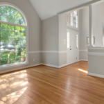 Bright living room with a large arched window, sunlit hardwood floors, light gray walls with white trim, and an open entryway leading to a kitchen/bar area.
