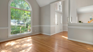 Bright living room with a large arched window, sunlit hardwood floors, light gray walls with white trim, and an open entryway leading to a kitchen/bar area.