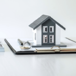 Small model house on a clipboard with papers, a pen, and keys, representing real estate paperwork.