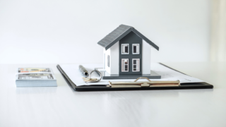 Small model house on a clipboard with papers, a pen, and keys, representing real estate paperwork.