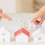 Two hands point at three white paper houses on a table, symbolizing comparing home options with charts in the background.