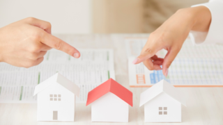 Two hands point at three white paper houses on a table, symbolizing comparing home options with charts in the background.