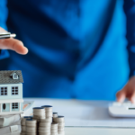 Businessperson in a blue suit points with a pen at a small model house resting on stacks of cash and coins, beside a calculator—illustrating real estate investment planning.