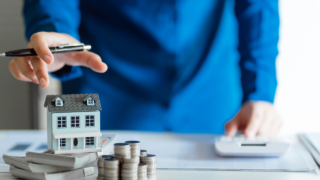 Businessperson in a blue suit points with a pen at a small model house resting on stacks of cash and coins, beside a calculator—illustrating real estate investment planning.